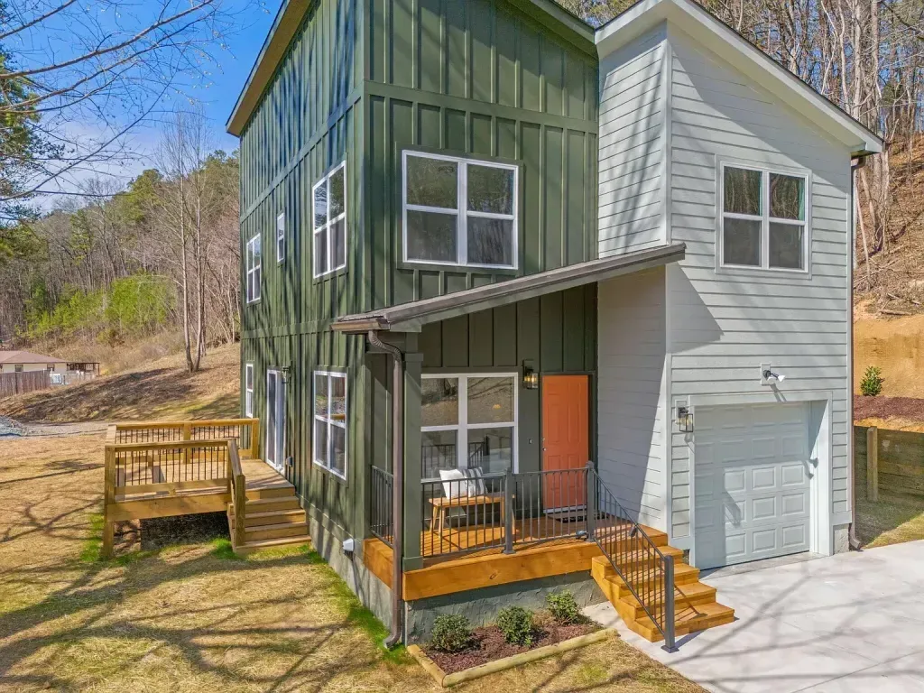 Two-story green board-and-batten home with a front porch and a wooden deck on a wooded hillside